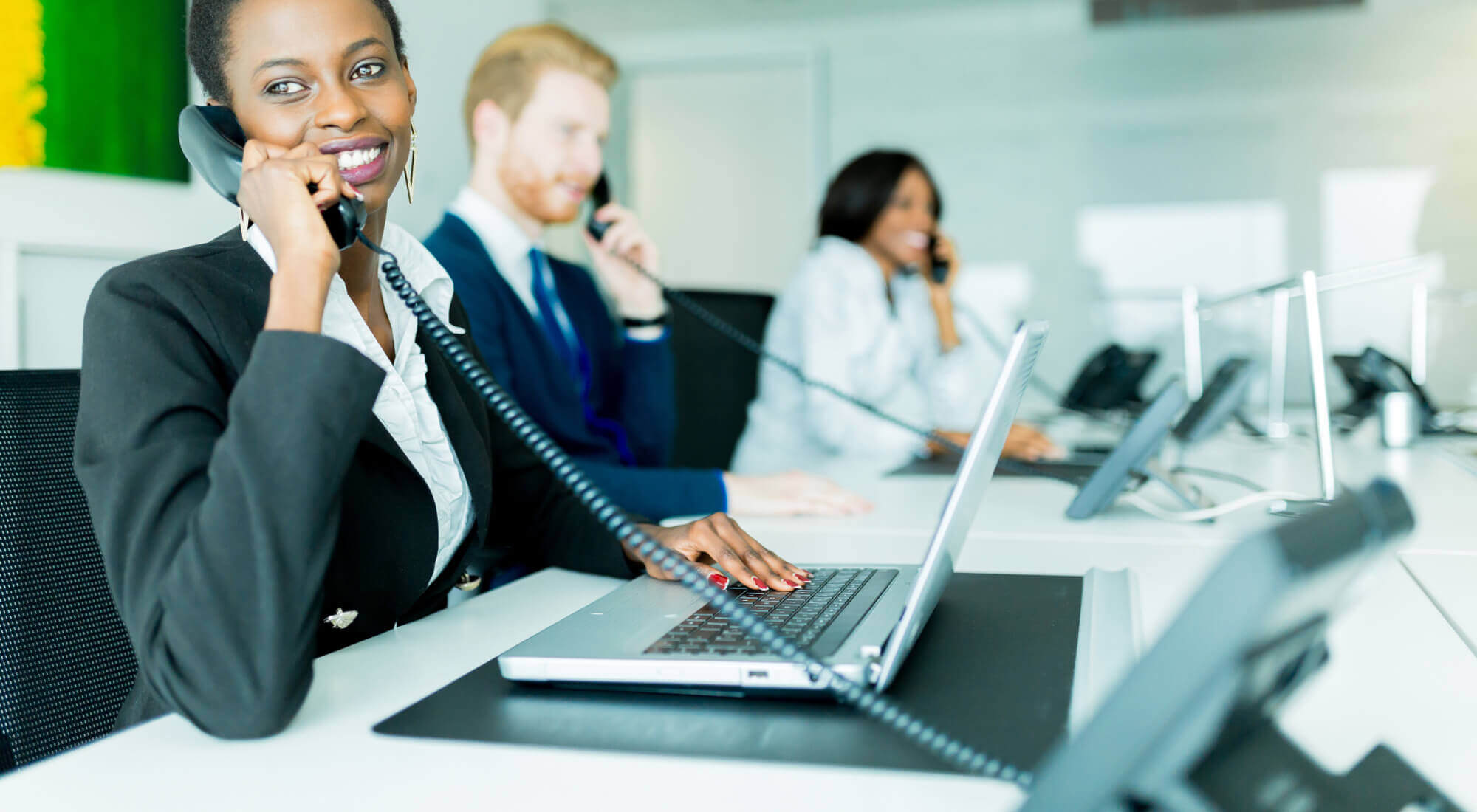 Smiling woman on desktop IP phone working on laptop in a call center