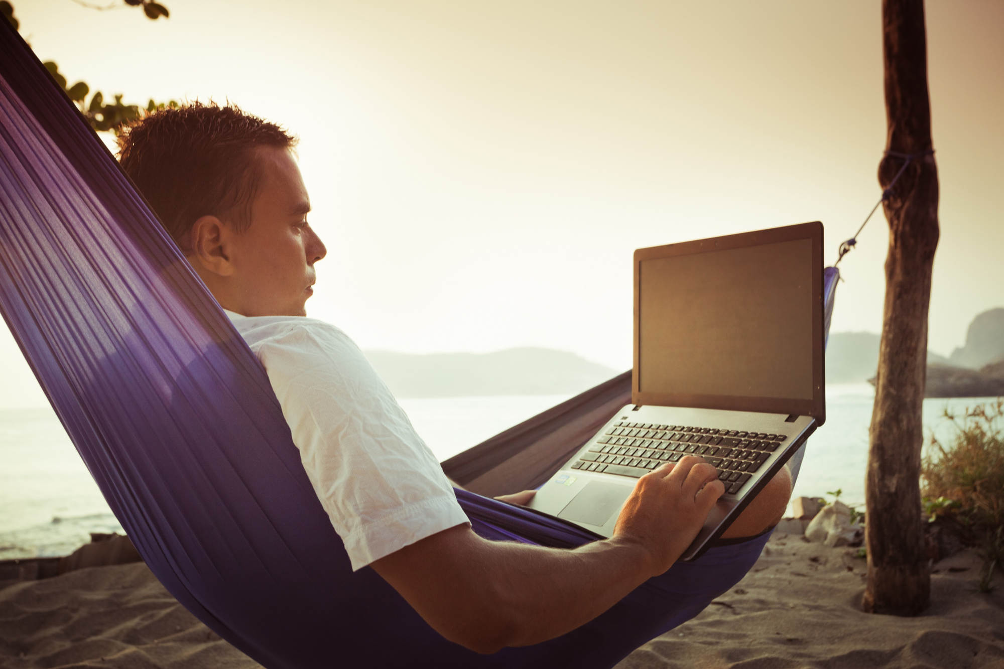 Man laying in purple hammock and using a laptop while at the beach